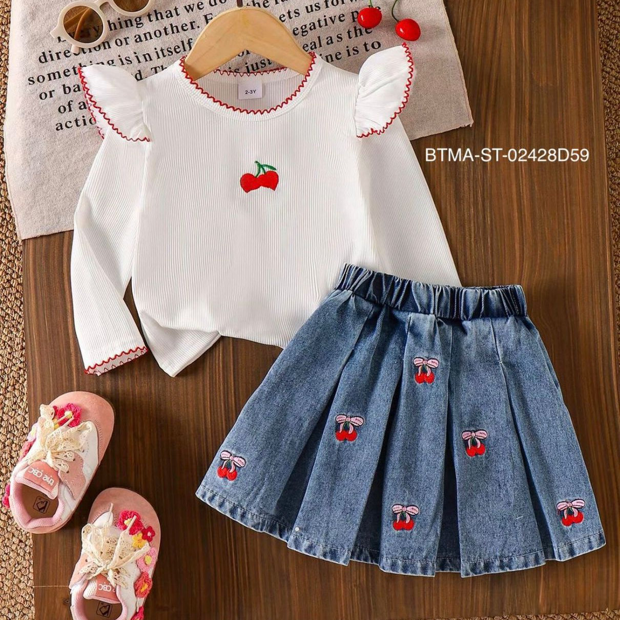 Children's outfit with white top and blue denim skirt featuring cherry designs, accompanied by pink sandals on a wooden surface.