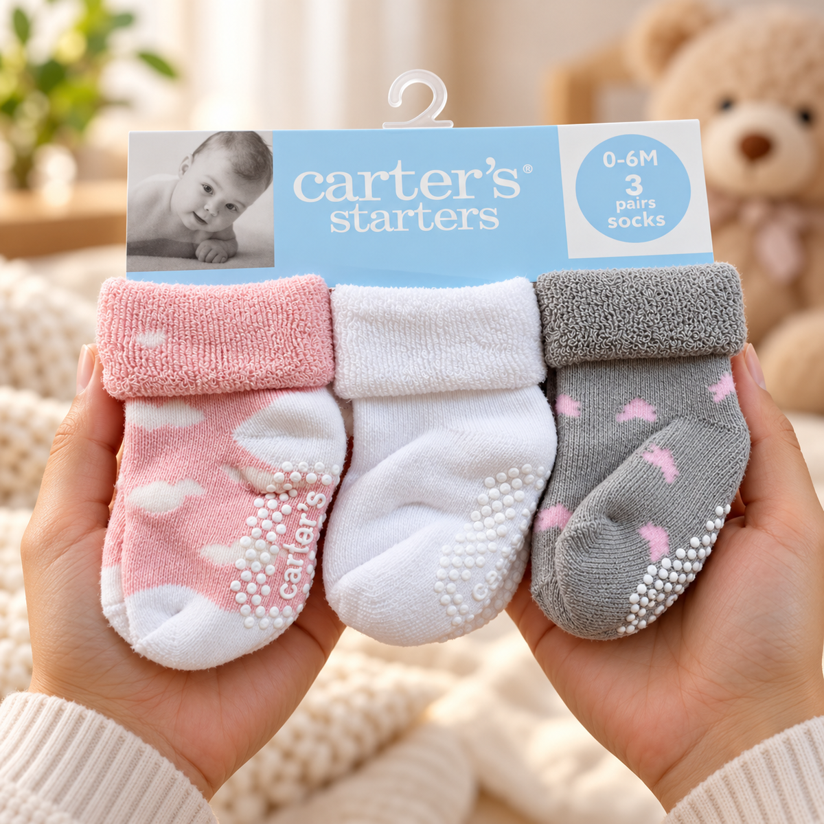 Carter’s starters 3-pair baby sock pack for 0-6 months, held in hands with Pink cloud-print, solid White and Grey Pink-heart socks showing White anti-slip sole grips.
