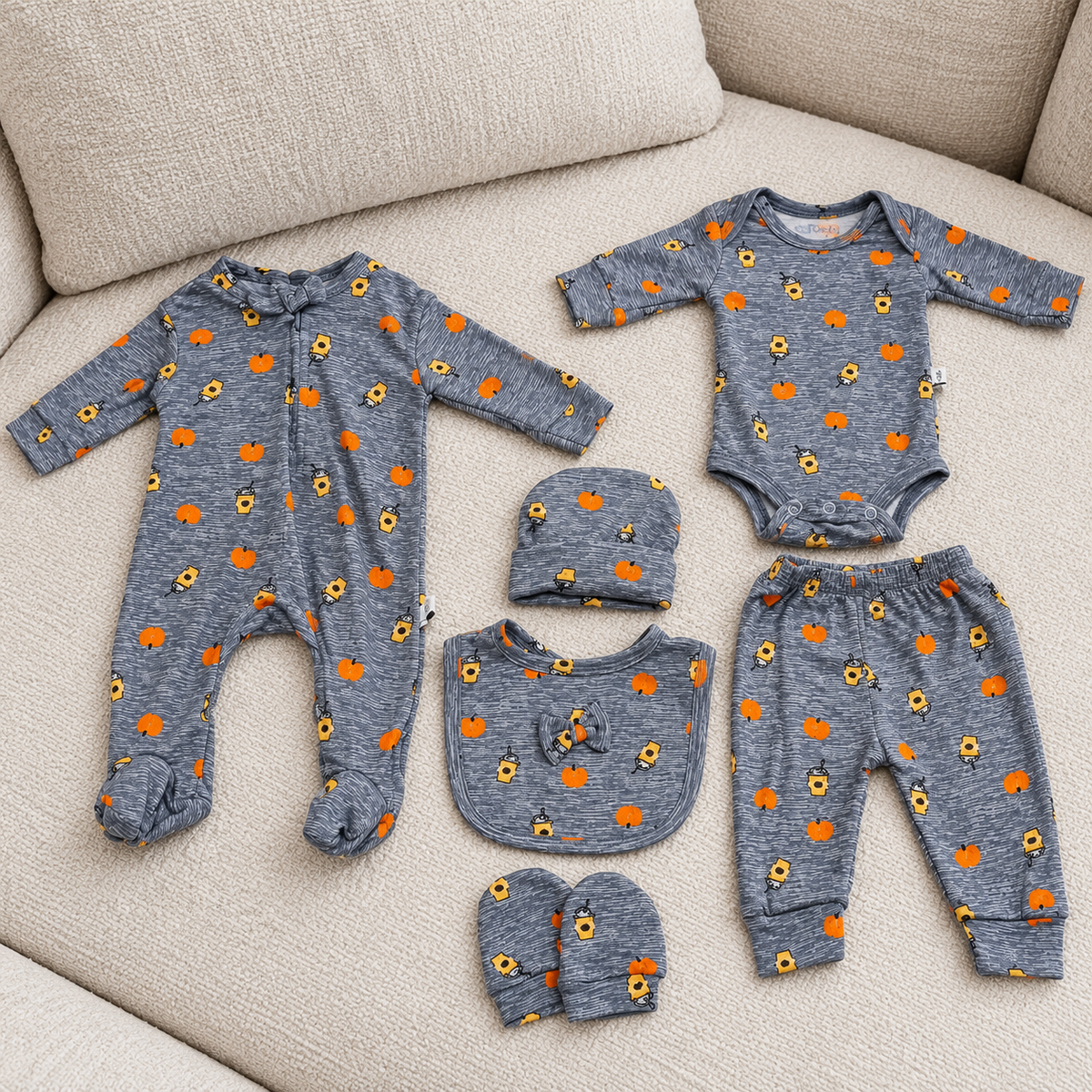 Orange pumpkin-print newborn 6pc starter set laid out on a cream sofa, with footed zip romper, long-sleeve bodysuit, trouser, cap, mittens and matching bib.
