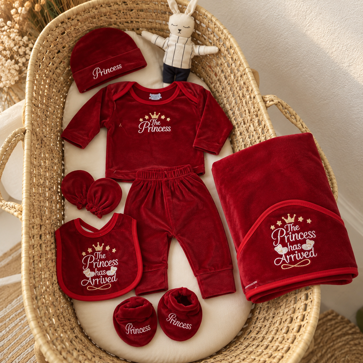 Red baby Princess velvet 8-piece starter set displayed in a wicker bassinet, featuring The Princess has Arrived embroidery across the top, bib and blanket.