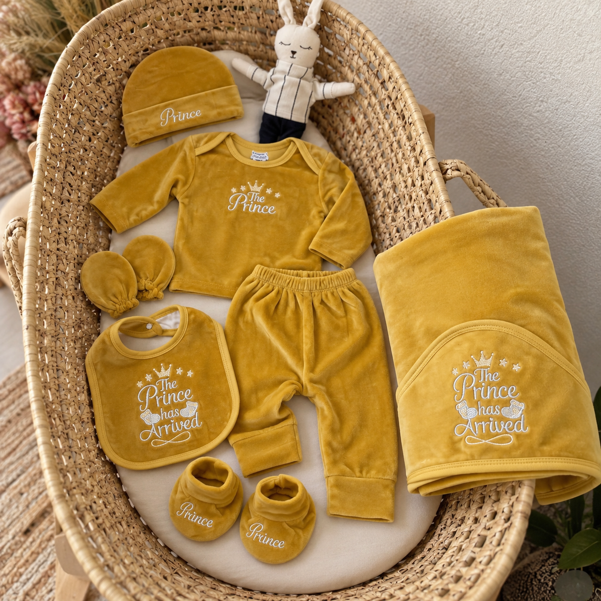 Yellow baby Prince velvet 8-piece starter set arranged in a wicker bassinet, showing the crown-print top, trouser, cap, mittens, bib, booties and matching blanket.
