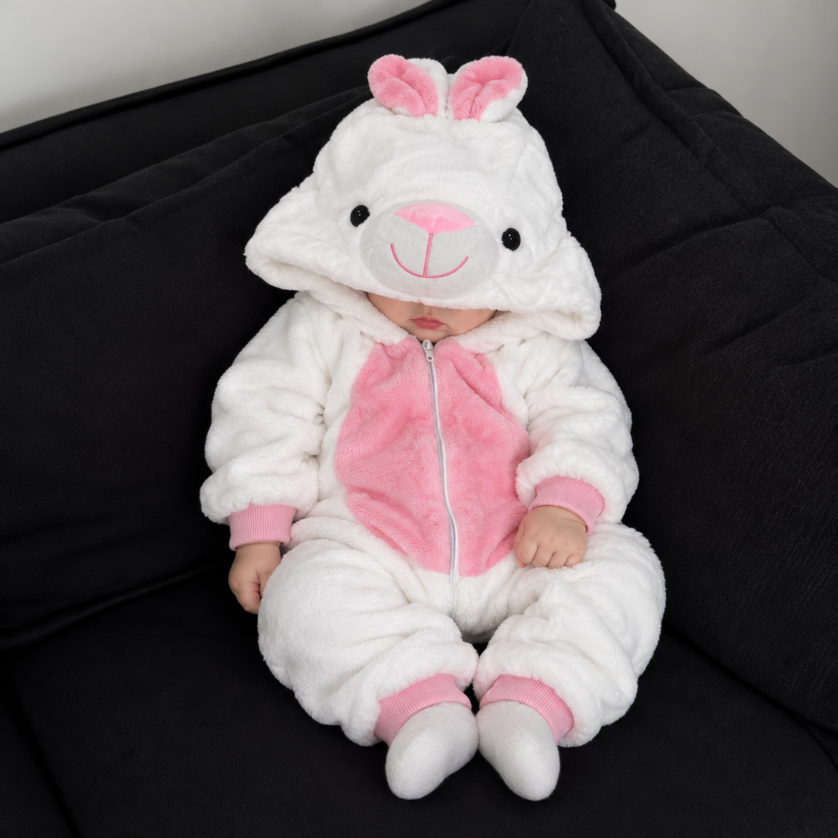 Baby seated on a dark sofa wearing the White and Pink bunny plush character romper with bunny-face hood, Pink tummy panel and ribbed cuffs.