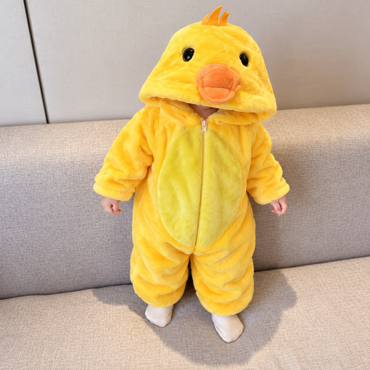 Baby wearing the Yellow chick-inspired plush hooded romper on a sofa, showing the raised Orange beak, small top crest, light Yellow tummy panel and front zip.