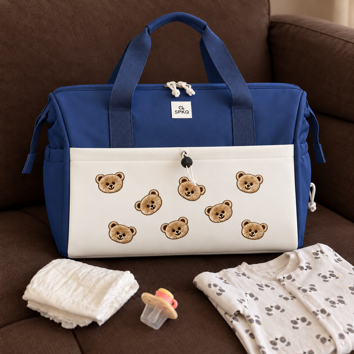 Blue easy carry mommy bag with a White teddy bear applique front pocket, styled on a sofa beside a nappy, pacifier and baby sleepsuit