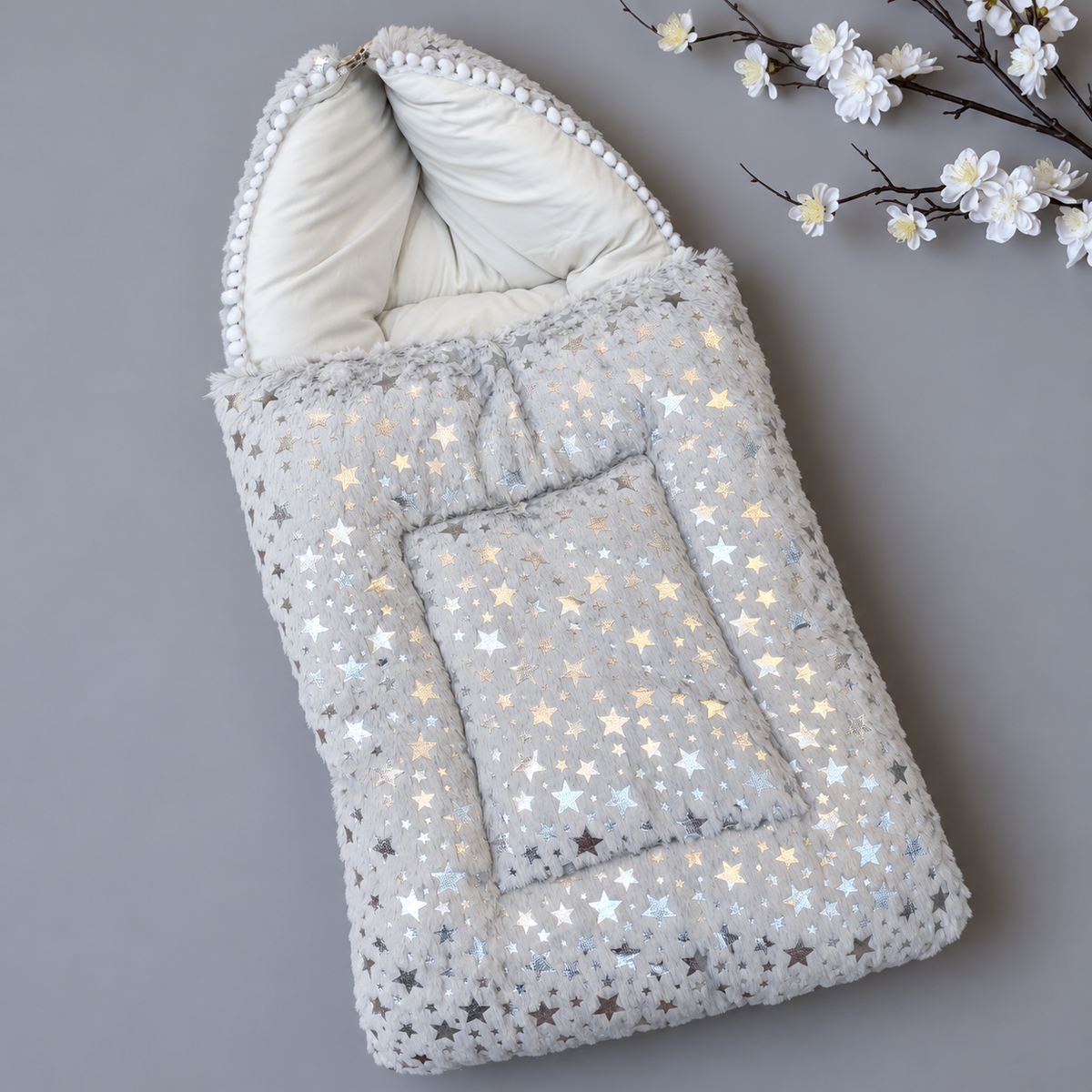 Grey baby shaggy carry nest shown in a diagonal flat lay, featuring metallic-look scattered stars, White hood lining and pom-pom-edged hood.