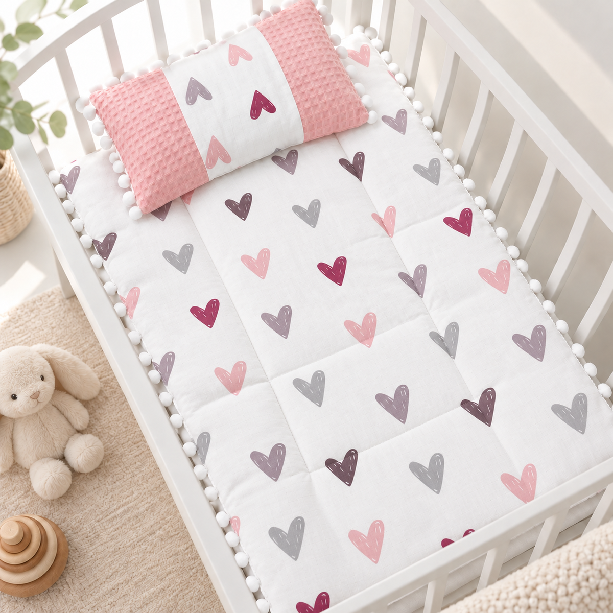 Pink heart printed baby sleeping sheet and matching Pink textured-side pillow, styled neatly in a White nursery cot with pom pom trim.