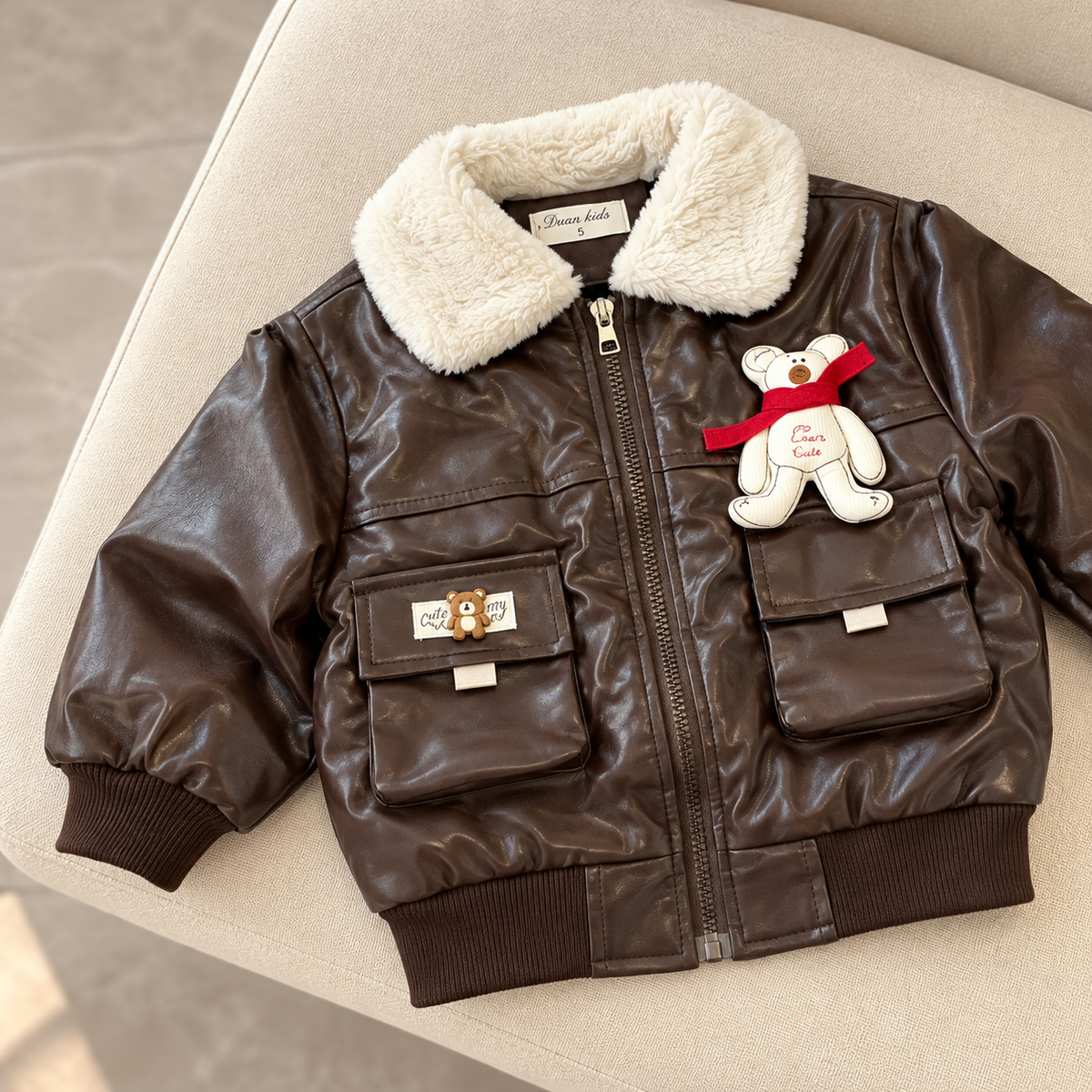 Brown kids leather-look winter jacket displayed flat on a beige chair, featuring a cream plush collar, zip front, teddy chest patch and two flap pockets.