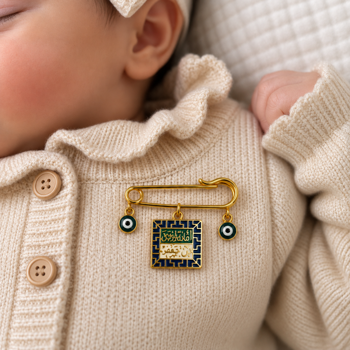 Gold-tone safety pin baby brooch on a Cream cardigan, with a square Arabic-script style pendant and two Green evil-eye beads.