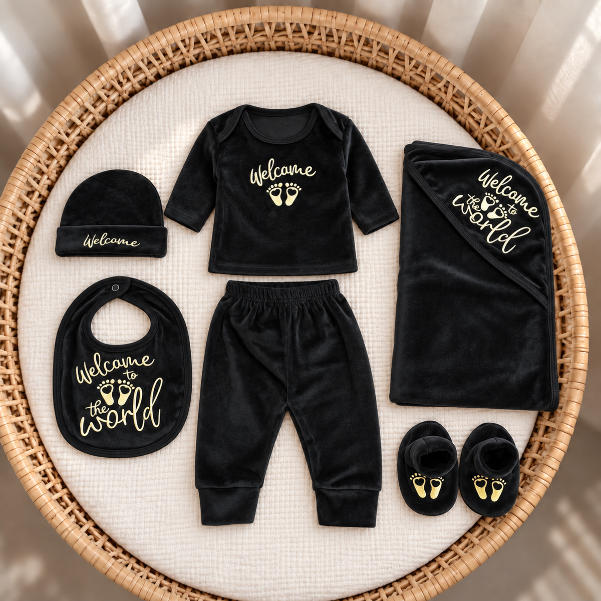 Black newborn 7-piece velvet starter set in a wicker basket, featuring gold-tone Welcome to the World lettering on the top, bib, blanket and booties.