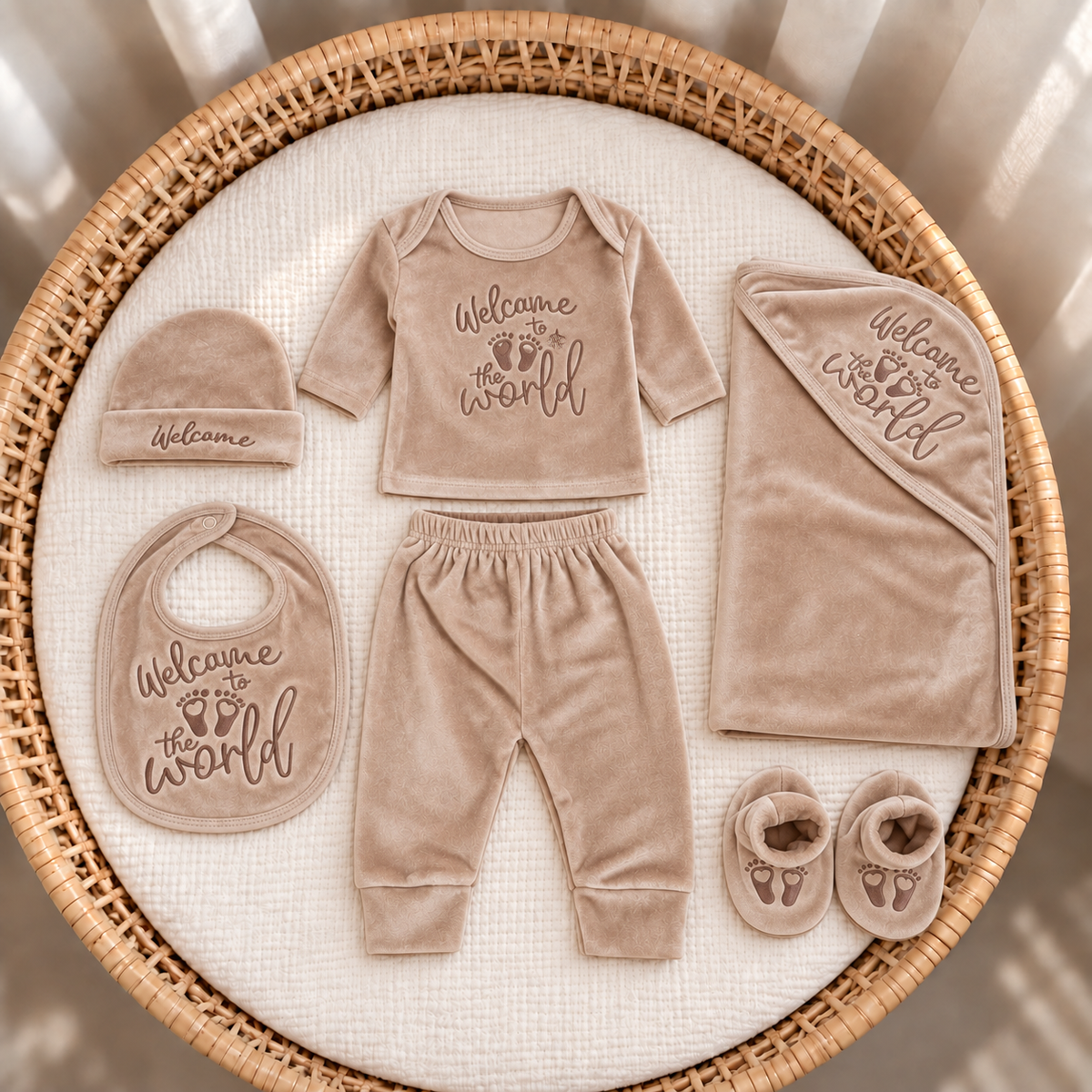 Brown newborn 7-piece velvet starter set displayed flat in a round wicker basket with matching Welcome to the World print and footprint-detail booties.