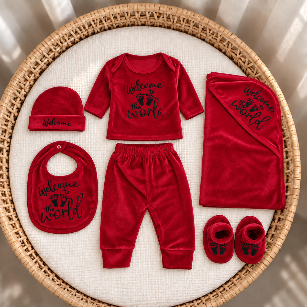 Red newborn 7-piece velvet starter set arranged in a wicker basket, showing Welcome to the World printed top, trouser, cap, bib, blanket and footprint booties.