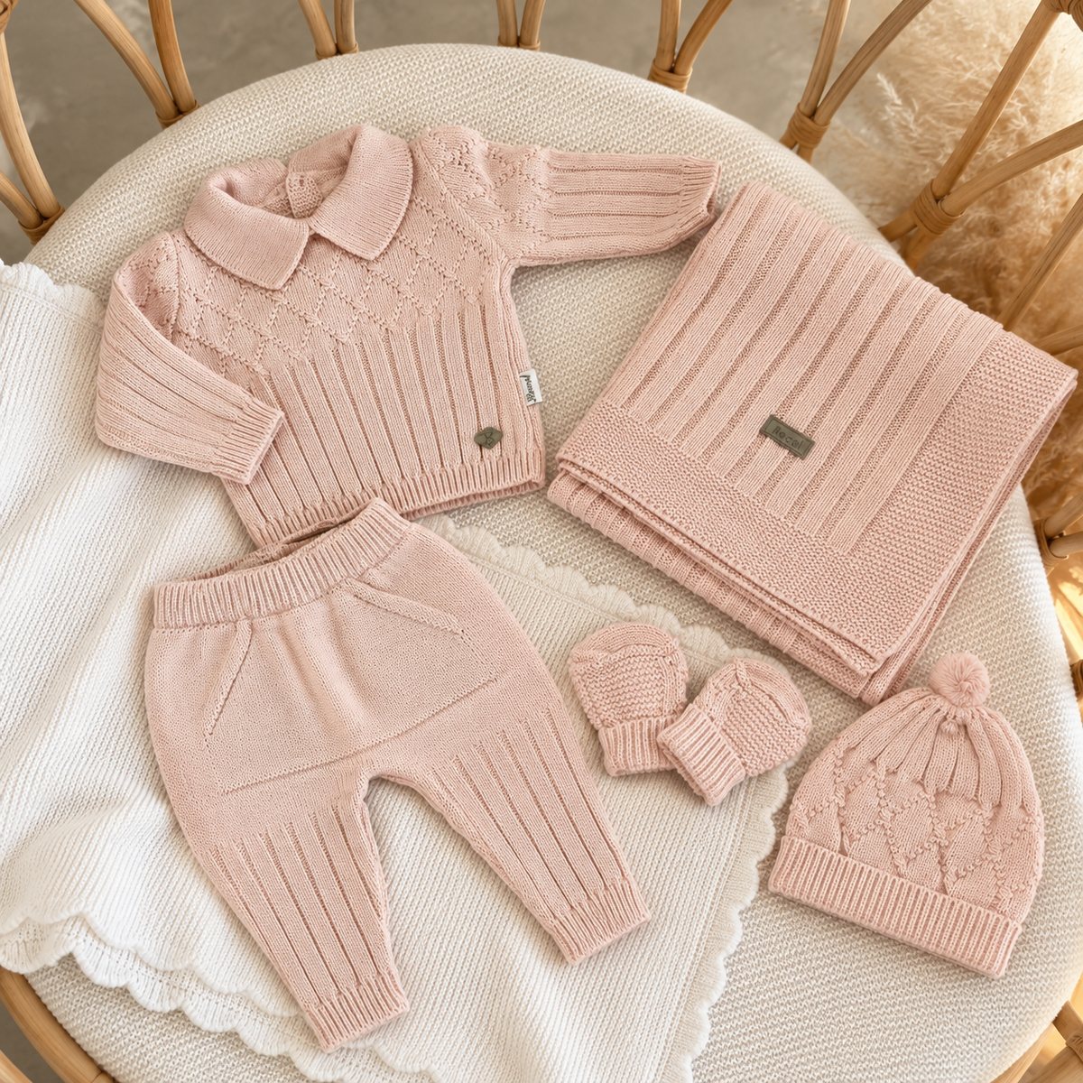 Light Pink newborn 5-piece knitted starter set arranged on a wicker chair, showing a collared cable-knit top, pocket trousers, blanket, pom-pom cap and mittens.