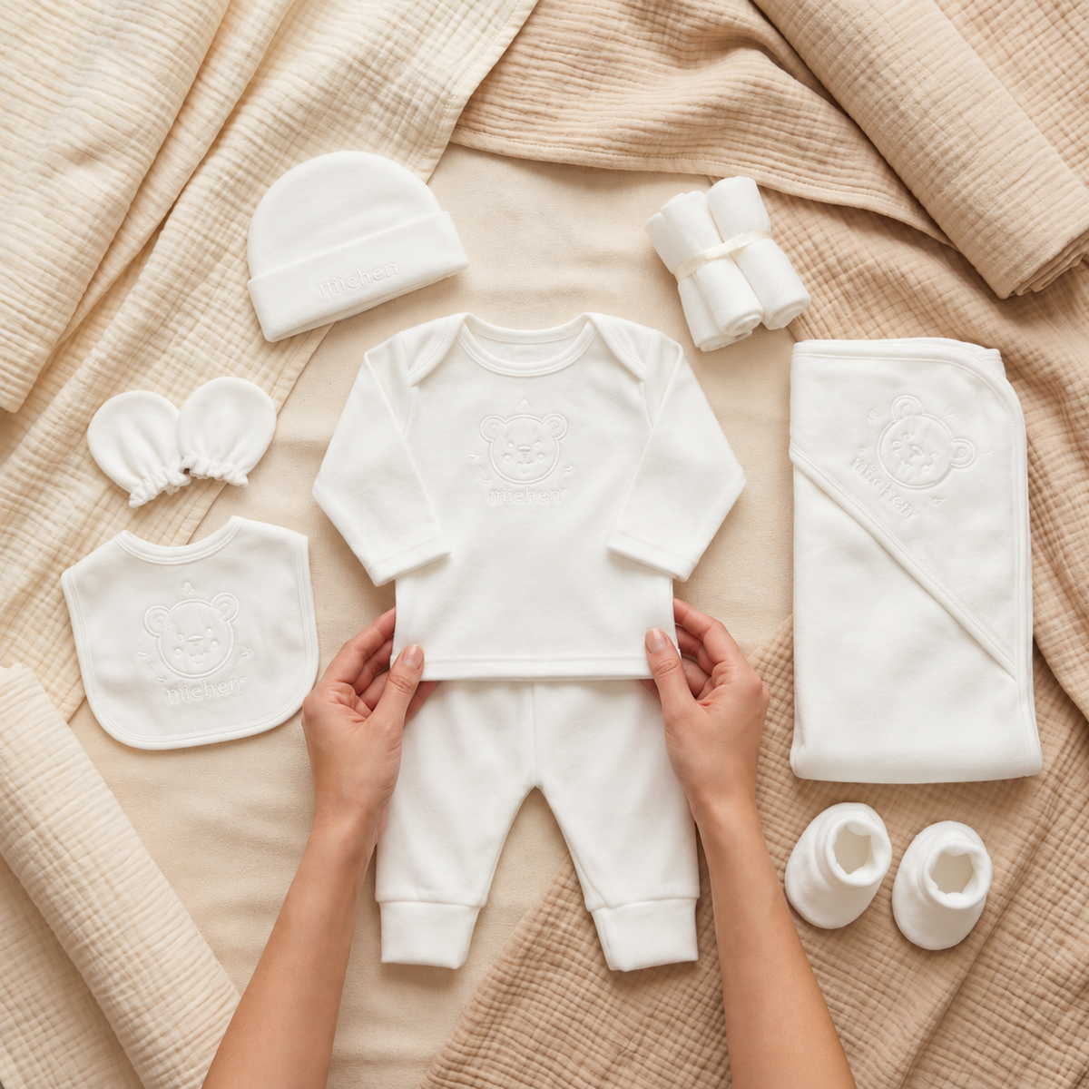 White newborn 11-piece starter set flat lay with teddy embroidered shirt, cuffed trousers, cap, bib, mittens, booties and matching wrap blanket.