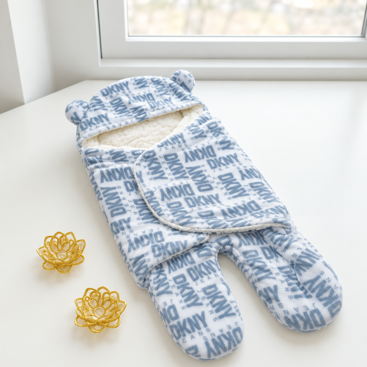 Light Blue baby footed swaddle displayed open by a window, with Blue DKNY-style all-over lettering, bear-ear hood and cream textured lining.