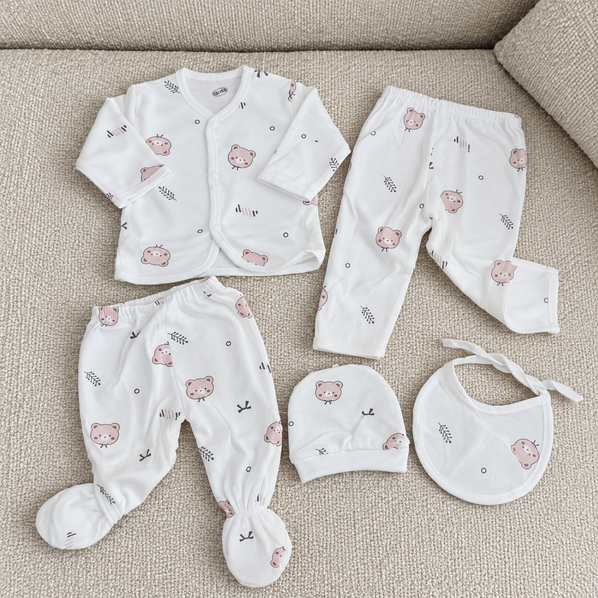 White baby bear print cotton 5-piece starter set flat lay on a textured sofa, showing the snap wrap top, regular trouser, footed trouser, cap and tie bib.