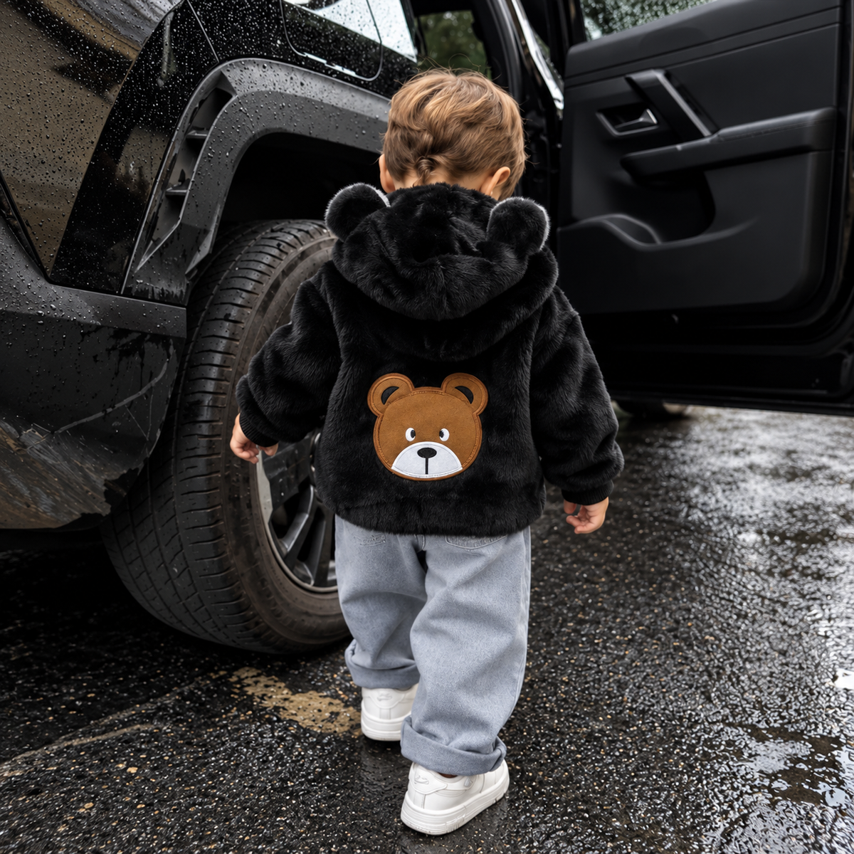 Child wearing the Black plush hooded jacket from the back beside a vehicle, showing the rounded bear-ear hood and large bear-face back patch.