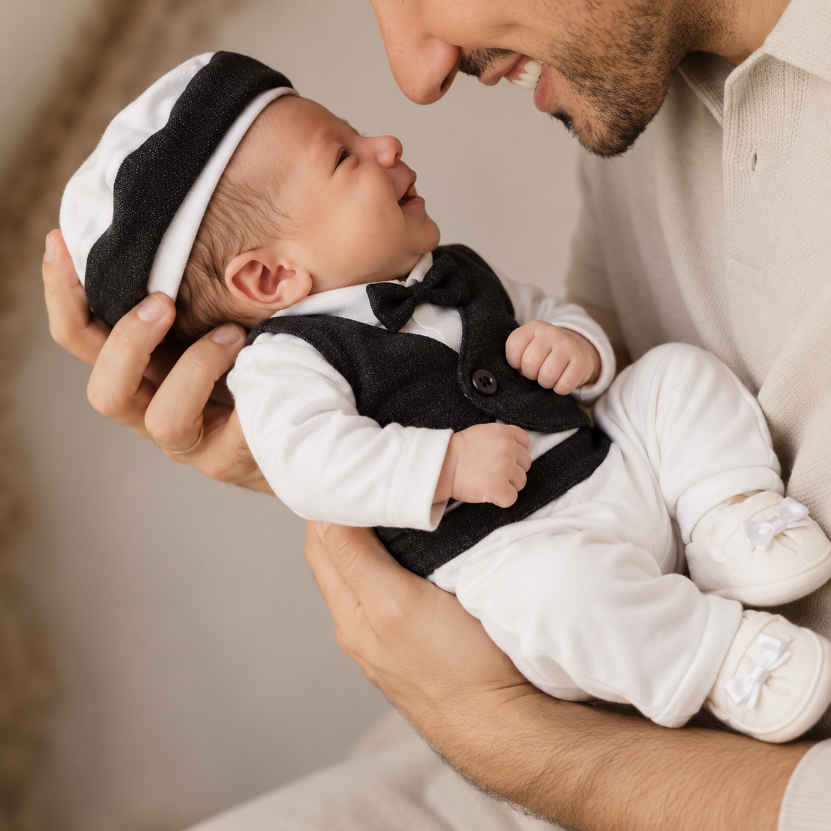 Newborn baby wearing the Black tuxedo-inspired starter set with White shirt, textured Black waistcoat, bow tie, cap and White bow-detail shoes while being held by an adult.