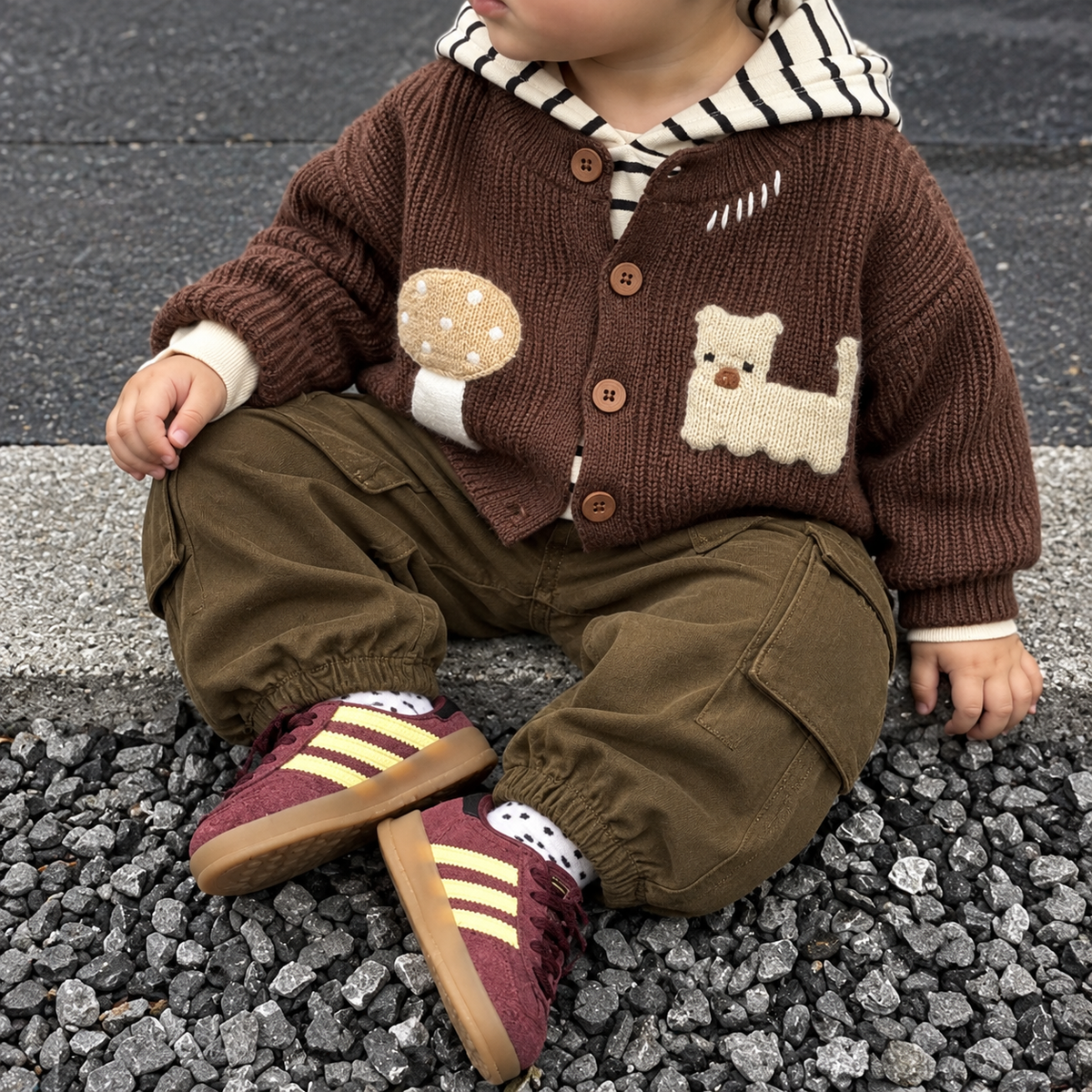 Toddler wearing a Brown textured knit-look button cardigan with bear and mushroom patches, striped hooded shirt and Olive Green cargo trousers on gravel.
