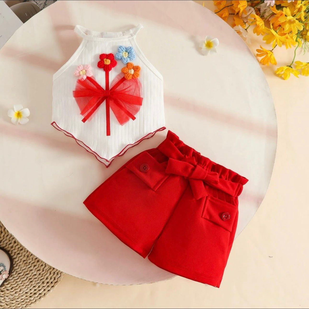 Children's outfit with white top and red shorts on a pink surface with flowers in the background