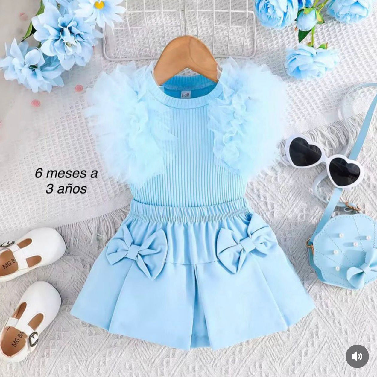 Light blue children's outfit with ruffles and bows on a textured surface with flowers and shoes.