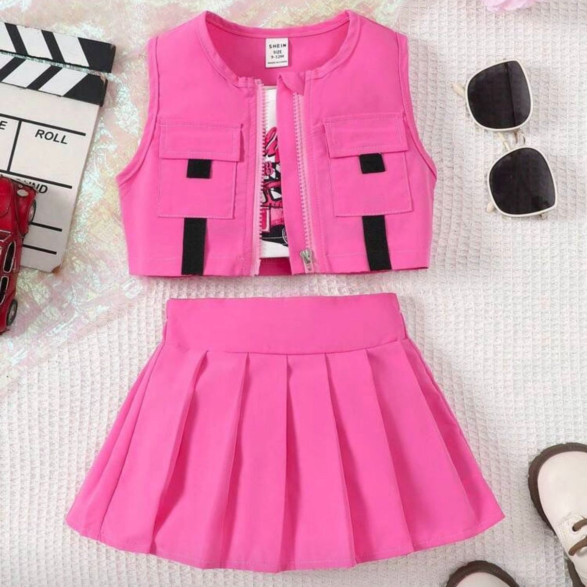 Pink vest and skirt set on a textured surface with flowers and clapperboard in the background.