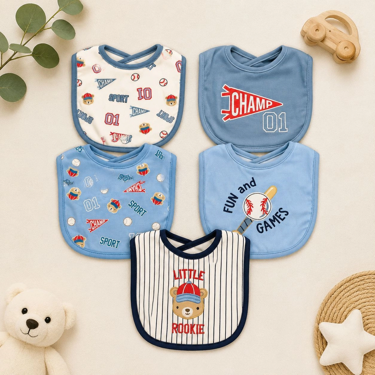 Five baby feeding bibs arranged on a cream backdrop, featuring Blue baseball prints, a CHAMP 01 graphic, Fun and Games bat artwork and a Little Rookie bear design.